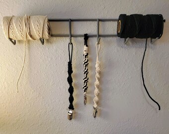 Pode incluir: Um rack de metal com quatro itens pendurados. Dois são rolos de corda, um é branco e o outro é preto. Os outros dois são prendedores de chupeta de crochê preto e branco.
