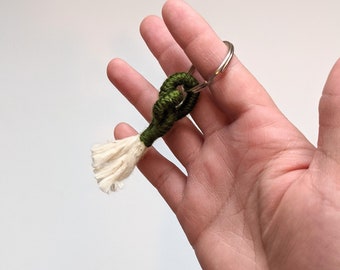 Pode incluir: Um chaveiro artesanal com um nó verde escuro e uma borla creme. Está preso a um anel de metal prateado. O design é simples e elegante, ideal para chaves ou acessórios.