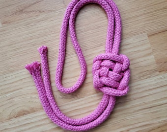 Pode incluir: Uma corda trançada rosa com um nó no centro. A corda está sobre uma superfície de madeira.