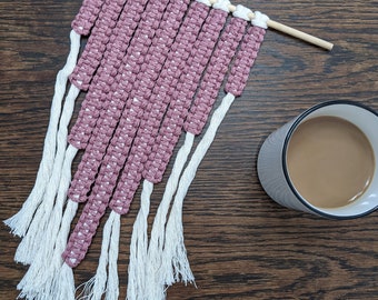 Pode incluir: Uma tapeçaria de macramé rosa e branca com uma vareta de madeira na parte superior e longas franjas brancas na parte inferior.