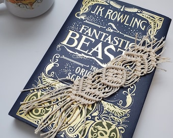 Pode incluir: Um livro de capa dura azul marinho com letras douradas que dizem "J.K. Rowling Fantastic Beasts The Original Screenplay". Um marcador de livro de macramé bege está preso ao livro.
