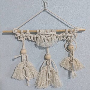 Puede incluir: Un tapiz de macramé de pared con tres figuras hechas de cuerda de fibra natural y cuentas de madera. Las figuras cuelgan de una varilla de madera.