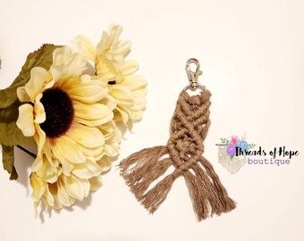 Pode incluir: Um chaveiro de macramé marrom com uma franja. O chaveiro está preso a um chaveiro prateado. O chaveiro é fotografado contra um fundo branco com um buquê de girassóis amarelos.