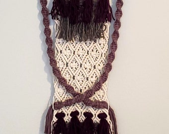 Pode incluir: Uma tapeçaria de parede em macramé com um padrão de losangos creme, borlas roxas escuras e cinzentas e detalhes em corda torcida. A decoração de parede é suspensa de um pino de madeira com uma corda para pendurar.