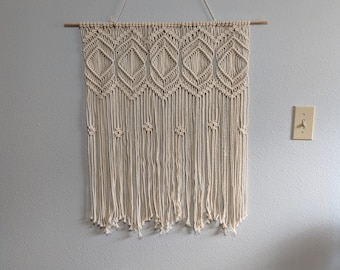 Pode incluir: Uma tapeçaria de macramé bege com um padrão de diamante e franjas. A tapeçaria está presa a uma vareta de madeira.