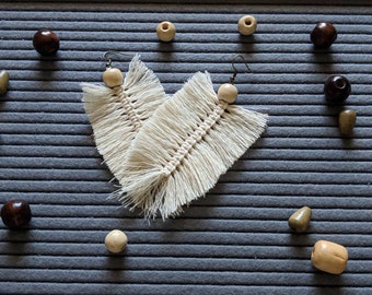 Pode incluir: Um par de brincos de pena de macramé branco com contas de madeira. Os brincos estão sobre uma superfície azul texturizada com contas de madeira espalhadas ao redor.