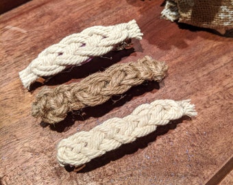 Pode incluir: Três presilhas de cabelo trançadas em tons de bege e castanho. As presilhas são feitas de um material semelhante a corda e estão dispostas sobre uma superfície de madeira. Cada presilha tem um fecho tipo barrette.