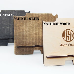 Puede incluir: Tres soportes de tel&eacute;fono de madera en diferentes acabados: tinte de &eacute;bano, tinte de nogal y madera natural. El soporte de madera natural tiene un monograma personalizado y el nombre "John Smith" grabado.