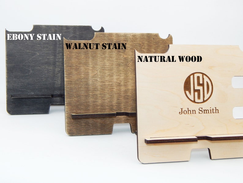 Puede incluir: Tres soportes de tel&eacute;fono de madera en diferentes acabados: tinte de &eacute;bano, tinte de nogal y madera natural. El soporte de madera natural tiene un monograma personalizado y el nombre "John Smith" grabado.