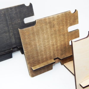 Puede incluir: Tres soportes de tel&eacute;fono de madera en diferentes acabados. El soporte de la izquierda es negro, el soporte del medio es marr&oacute;n y el soporte de la derecha es de madera natural con un monograma "JS" en un c&iacute;rculo. Los soportes tienen una ranura para un tel&eacute;fono y una plataforma elevada para cargar.