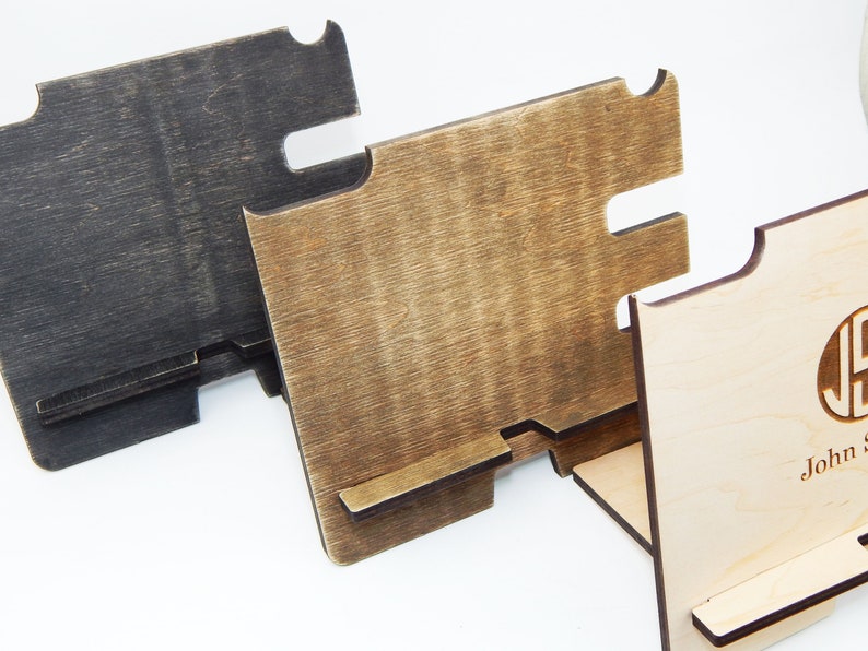 Puede incluir: Tres soportes de tel&eacute;fono de madera en diferentes acabados. El soporte de la izquierda es negro, el soporte del medio es marr&oacute;n y el soporte de la derecha es de madera natural con un monograma "JS" en un c&iacute;rculo. Los soportes tienen una ranura para un tel&eacute;fono y una plataforma elevada para cargar.