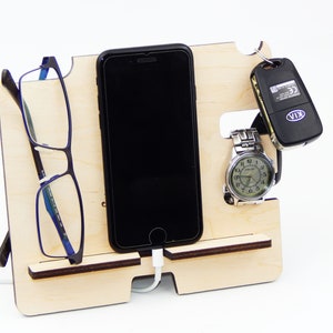 Puede incluir: Un soporte de tel&eacute;fono de madera con ranuras para un tel&eacute;fono, gafas, un reloj y llaves. El soporte est&aacute; hecho de madera clara y tiene un acabado natural. El tel&eacute;fono es un iPhone negro con un cable de carga enchufado. Las gafas son azules y negras. El reloj es plateado con una esfera negra. Las llaves son negras con un logotipo KIA plateado.