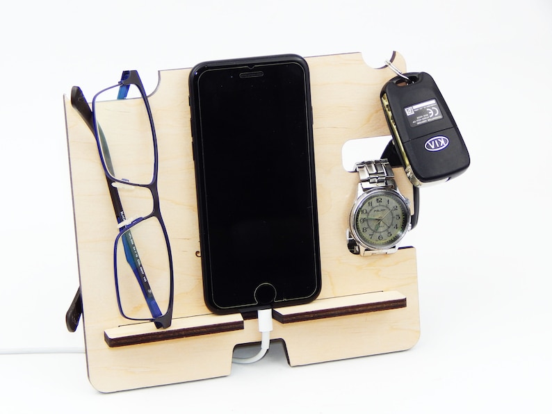 Puede incluir: Un soporte de tel&eacute;fono de madera con ranuras para un tel&eacute;fono, gafas, un reloj y llaves. El soporte est&aacute; hecho de madera clara y tiene un acabado natural. El tel&eacute;fono es un iPhone negro con un cable de carga enchufado. Las gafas son azules y negras. El reloj es plateado con una esfera negra. Las llaves son negras con un logotipo KIA plateado.