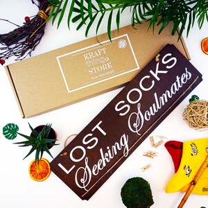 May include: A brown wooden sign with white text that reads "Lost Socks Seeking Soulmates". The sign is hanging from a string and is surrounded by various items, including a small potted plant, a wicker ball, and a yellow sock with a red heel.