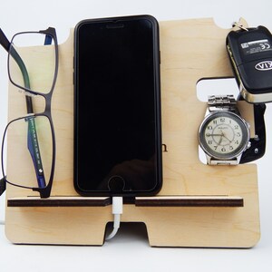 Puede incluir: Un organizador de madera con ranuras para un tel&eacute;fono, gafas, un reloj y llaves. El tel&eacute;fono se est&aacute; cargando con un cable blanco. El reloj es plateado con una esfera negra y agujas negras. Las llaves son negras con un logotipo KIA plateado. El organizador est&aacute; hecho de madera clara.