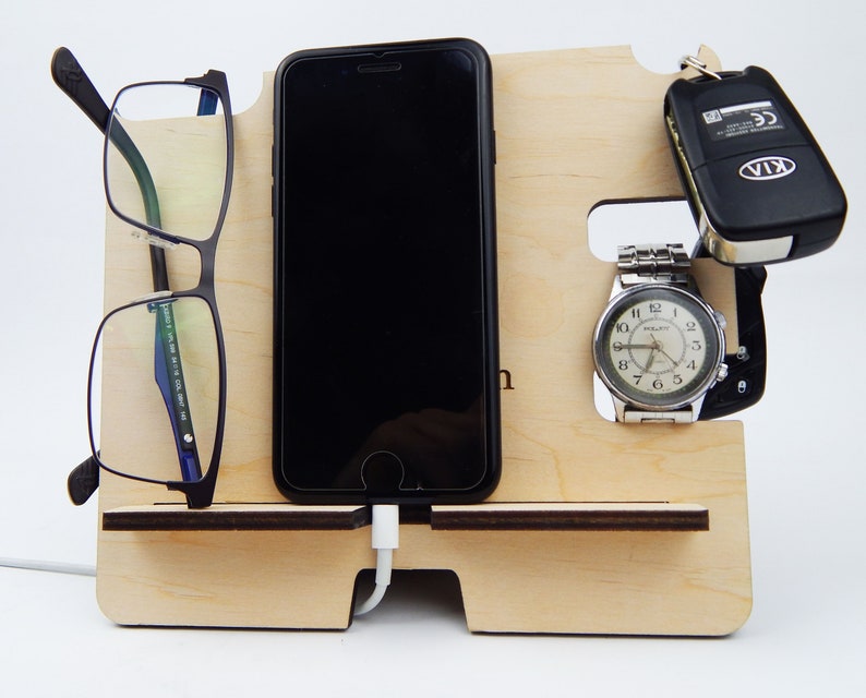Puede incluir: Un organizador de madera con ranuras para un tel&eacute;fono, gafas, un reloj y llaves. El tel&eacute;fono se est&aacute; cargando con un cable blanco. El reloj es plateado con una esfera negra y agujas negras. Las llaves son negras con un logotipo KIA plateado. El organizador est&aacute; hecho de madera clara.