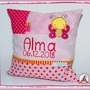 May include: Pink and yellow personalized pillow with a butterfly design and the name "Alma" and the date "06.12.2018" embroidered on it.