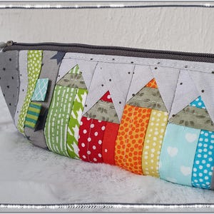 Könnte beinhalten: Ein gefüttertes Federmäppchen mit grauem Reißverschluss und einem farbenfrohen Bleistiftmuster. Das Etui hat eine Vielzahl von Stoffen, darunter Punkte, Streifen und Sterne. Das Bleistiftmuster umfasst rote, orangefarbene, gelbe, grüne und blaue Bleistifte.