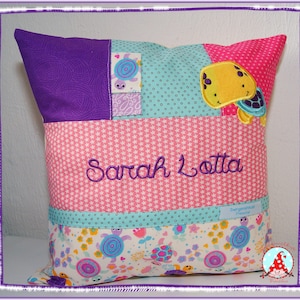May include: A pink and purple pillow with a patchwork design. The pillow has a pocket on the front with a turquoise fabric and a yellow turtle applique. The pillow is embroidered with the name "Sarah Lotta".