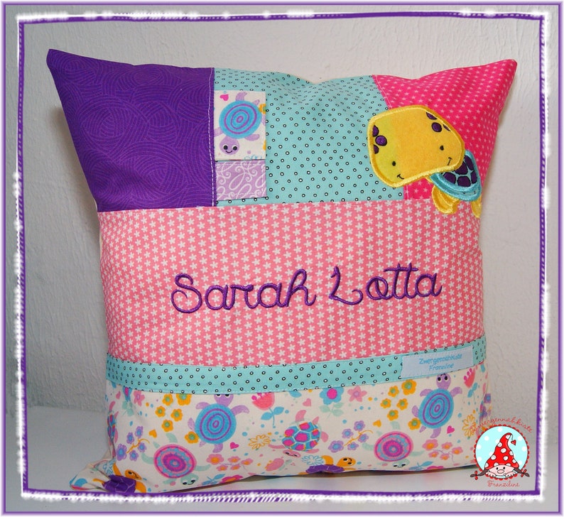 May include: A pink and purple pillow with a patchwork design. The pillow has a pocket on the front with a turquoise fabric and a yellow turtle applique. The pillow is embroidered with the name "Sarah Lotta".