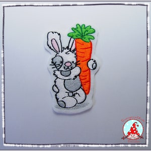 Könnte beinhalten: Gestickter Aufnäher eines grauen Kaninchens, das eine große orange Karotte mit grünen Blättern hält.