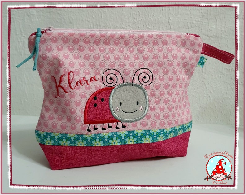 May include: Pink zippered pouch with a ladybug applique and the name "Klara" embroidered in red. The pouch features a pink polka dot pattern, a teal floral trim, and a red base. A teal pull cord and a red fabric loop are attached to the zipper.