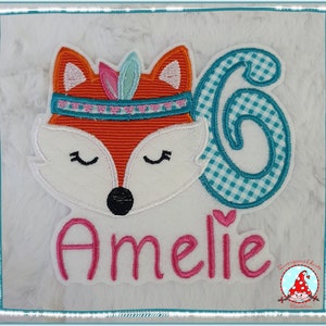 Könnte beinhalten: Ein weißer Filz-Patch mit einem bunten gestickten Fuchs, der ein Stirnband mit Federn trägt, einer türkisfarbenen Nummer 6 und dem Namen "Ameli" in rosa gestickt.