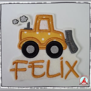 Könnte beinhalten: Gelbe Filzapplikation eines Spielzeugbulldozers mit weißen Punkten und dem Namen Felix in goldenen Filzbuchstaben.