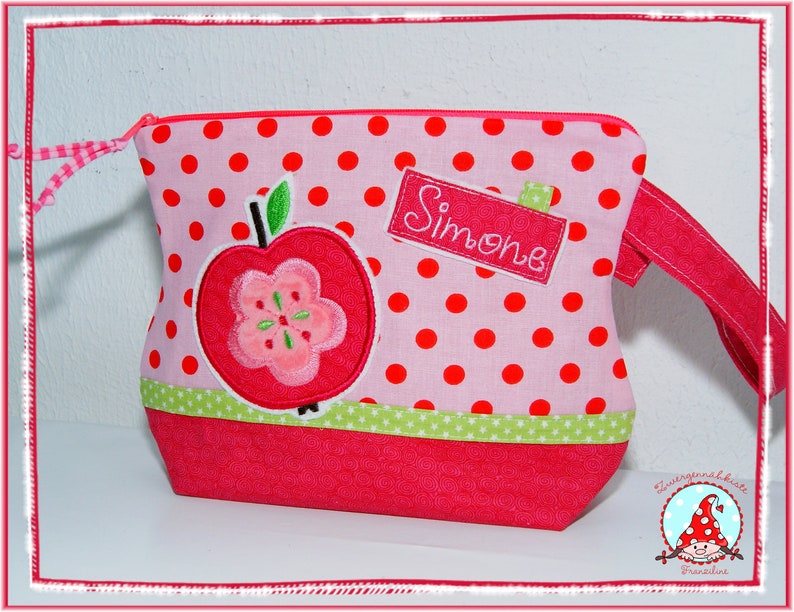 May include: Pink polka dot bag with a red apple applique and the name "Simone" embroidered on a label. The bag features a red zipper, a green and white star trim, and a red strap. The bag is designed with a playful, whimsical aesthetic.