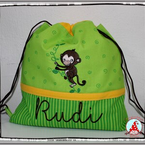 Könnte beinhalten: Grüne Kordelzugtasche mit gelbem Streifen und einem Affenmotiv. Die Tasche hat den Namen "Rudi" auf der Vorderseite gestickt.