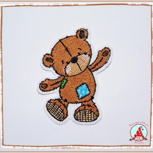 Könnte beinhalten: Ein brauner Teddybär mit einem blauen Flicken auf seinem Bauch. Der Bär steht mit erhobenen Armen und trägt eine grüne Schleife. Der Bär ist auf einem weißen Hintergrund gestickt.