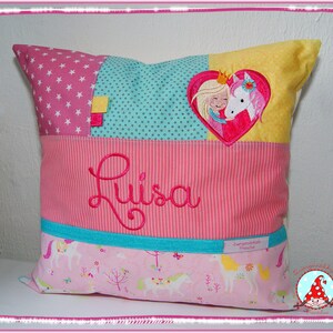 Könnte beinhalten: Ein rosa-blaues Kissen mit einem Einhorn- und Prinzessinnen-Design. Das Kissen hat eine Tasche mit dem Namen "Luisa" bestickt.