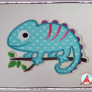 Könnte beinhalten: Ein blauer Chamäleon mit weißen Herzen und rosa Streifen sitzt auf einem braunen Ast mit grünen Blättern. Das Chamäleon hat ein großes weißes Auge.