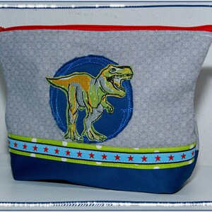 May include: Gray zippered pouch with a dinosaur applique. The pouch features a blue and green dinosaur design on a blue circle, a red zipper, and a blue and red star pattern on the bottom. The pouch is approximately 8 inches wide and 6 inches tall.