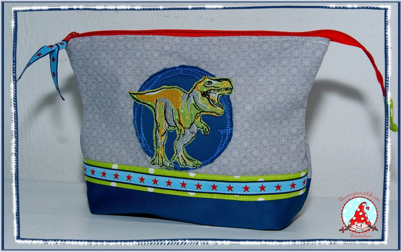May include: Gray zippered pouch with a dinosaur applique. The pouch features a blue and green dinosaur design on a blue circle, a red zipper, and a blue and red star pattern on the bottom. The pouch is approximately 8 inches wide and 6 inches tall.
