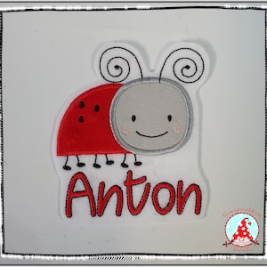 Könnte beinhalten: Rote und graue gestickte Marienkäfer mit schwarzen Beinen und weißen Antennen. Der Name Anton ist in Rot unter dem Marienkäfer gestickt.