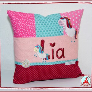 May include: A pink and red pillow with a patchwork design featuring a unicorn and the name "Lia" embroidered on it.