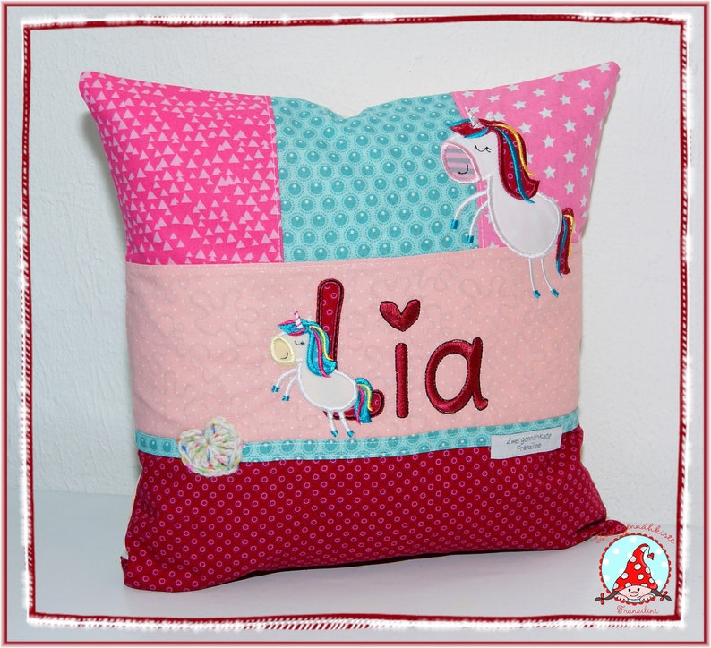 May include: A pink and red pillow with a patchwork design featuring a unicorn and the name "Lia" embroidered on it.