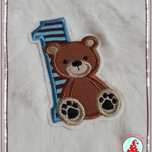 Könnte beinhalten: Ein brauner Teddybär-Applikation mit schwarzen Pfotenabdrücken sitzt auf einer blau-weiß gestreiften Nummer eins. Die Applikation ist von einem weißen Rand umgeben.