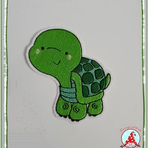 Könnte beinhalten: Grüne gestickte Schildkröten-Applikation mit einem lächelnden Gesicht und einem Panzer mit Karomuster. Die Schildkröte befindet sich auf einem weißen Hintergrund.