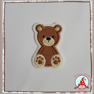 Könnte beinhalten: Ein braunes Teddybär-Applikationsmotiv mit weißen Pfoten und einem weißen Umriss. Der Bär sitzt mit seinen Pfoten vor sich.