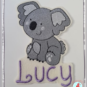 Könnte beinhalten: Ein gestickter grauer Koala, der mit seinen Pfoten vor sich sitzt. Der Name "Lucy" ist in Lila unter dem Koala gestickt.