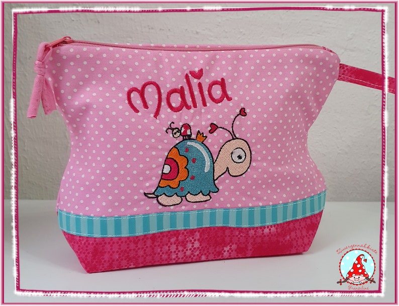 May include: Pink polka-dot cosmetic bag with the name "Malia" embroidered in red. Features a cartoon snail with a blue shell and orange accents. The bag has a turquoise stripe and a pink zipper.