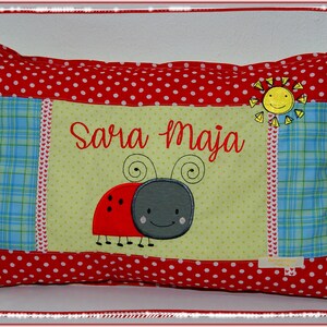 Könnte beinhalten: Ein rotes und grünes Kissen mit weißen Punkten. Das Kissen hat eine gelbe Sonne, einen Marienkäfer und den Namen "Sara Maja" gestickt.