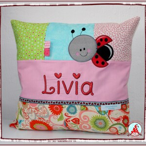Könnte beinhalten: Ein quadratisches Dekokissen mit Patchwork-Design aus grünen Punkten, hellblauem Stoff und einer Marienkäfer-Applikation. Das mittlere rosa Feld zeigt den in Rot gestickten Namen Livia. Der untere Bereich ist mit einem Blumenmuster versehen.