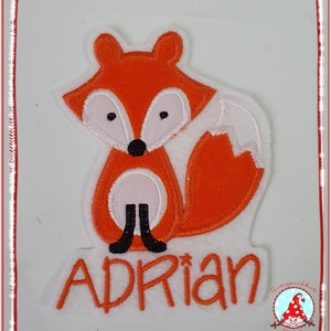 Könnte beinhalten: Orange und weiß gestickter Fuchs-Applikation mit dem Namen "Adrian" in orangefarbener Stickerei auf weißem Hintergrund.