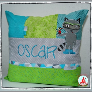 Könnte beinhalten: Ein grünes und blaues Kissen mit einer Waschbär- und Vogel-Applikation. Das Kissen hat eine graue Tasche mit dem Namen "Oscar" bestickt.