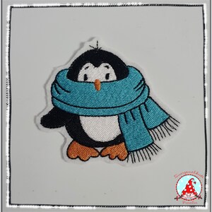 Könnte beinhalten: Gestickter Pinguin mit blauem Schal. Der Pinguin steht auf einem weißen Hintergrund.