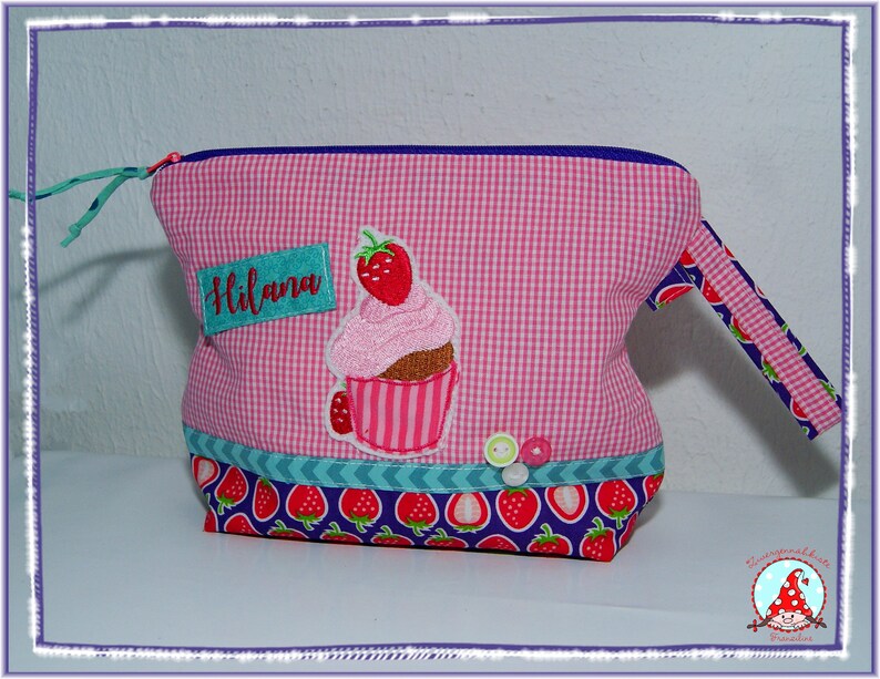 May include: Pink gingham fabric bag with a cupcake applique and the name "Hilana". The bag features a strawberry pattern on the bottom and handle, with a blue zipper and trim. Decorative buttons are sewn on the front.