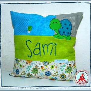 May include: A green and blue embroidered turtle is on a green and blue patchwork pillow with the name Sami embroidered on it. The pillow has a white background with a small turtle pattern.